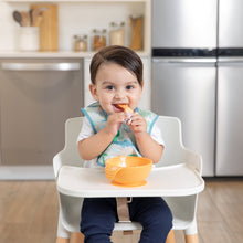 Load image into Gallery viewer, A dark-haired baby in a Bumkins Starter Bib 2 Pack: Dinosaurs &amp; Blue Tropic smiles in a high chair while eating from an orange bowl.
