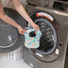 Load image into Gallery viewer, A person holds a Bumkins Starter Bib 2 Pack: Dinosaurs &amp; Blue Tropic in front of an open front-loading washing machine.

