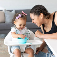 Load image into Gallery viewer, A woman smiles at a toddler in a high chair wearing a Bumkins Starter Bib 2 Pack: Floral &amp; Lace with a blue bowl on the tray.
