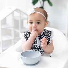 Load image into Gallery viewer, A baby girl in a black &amp; white bib and bow chews a Bumkins Silicone Dipping Spoons 3 Pack: Taffy near a gray bowl on a high chair tray.
