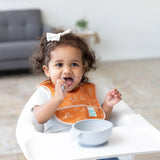 A toddler with curly hair wears an orange Bumkins Starter Bib 2 Pack: Sunshine and Grounded, eating with a spoon in a high chair.