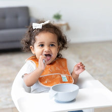 Load image into Gallery viewer, A toddler with curly hair wears an orange Bumkins Starter Bib 2 Pack: Sunshine and Grounded, eating with a spoon in a high chair.
