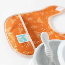 Load image into Gallery viewer, Close-up of an orange Bumkins Starter Bib 2 Pack: Sunshine and Grounded beside a gray baby bowl and spoon on a white background.
