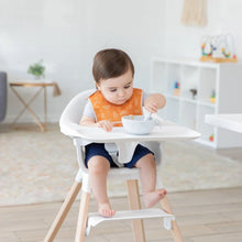 Load image into Gallery viewer, In a bright room, a toddler in a high chair uses a spoon to eat while wearing an orange Bumkins Starter Bib 2 Pack: Sunshine and Grounded.
