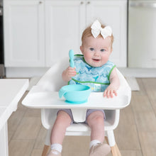 Load image into Gallery viewer, A baby sits in a high chair, wearing a Bumkins Starter Bib 2 Pack: Ocean Life &amp; Whale Tail and holding a blue spoon over a blue bowl.
