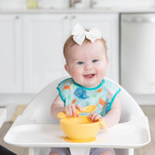 Load image into Gallery viewer, A baby in a high hair smiles, wearing a Bumkins Starter Bib 2 Pack: Ocean Life &amp; Whale Tail while holding a yellow bowl and spoon.
