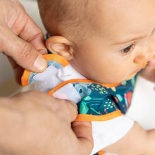 Load image into Gallery viewer, Adult hands fasten the hook-and-loop closure of a Bumkins Starter Bib 2 Pack: Jungle &amp; Animal Prints around a baby's neck.

