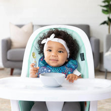 Load image into Gallery viewer, A baby in a high chair wears a Bumkins Starter Bib 2 Pack: Jungle &amp; Animal Prints, holding a gray spoon near a gray bowl.
