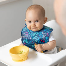 Load image into Gallery viewer, A baby in a high chair wears a Bumkins Starter Bib 2 Pack: Jungle &amp; Animal Prints, smiling and holding a spoon with a yellow bowl nearby.

