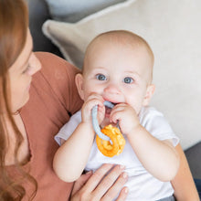 Load image into Gallery viewer, A baby with big blue eyes chews on a Bumkins Silicone Teething Charms: Orange, offering teething relief, held by a red-haired woman.
