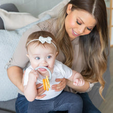 Load image into Gallery viewer, On a cozy couch, a woman smiles as her baby enjoys chewing on Bumkins Silicone Teething Charms: Orange while sporting a bow headband.
