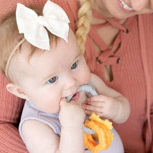 Load image into Gallery viewer, A baby with a white bow chews on a Bumkins Silicone Teething Charms: Orange, held by someone in a peach sweater.
