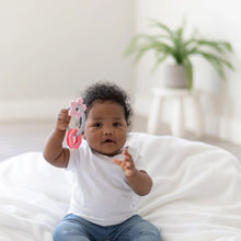 Load image into Gallery viewer, A baby in a white shirt and jeans sits on a white blanket, holding a Bumkins Silicone Teething Charms: Pink, with a potted plant behind.
