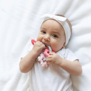 A baby in white outfit with a headband chews a Bumkins Silicone Teething Charms: Pink while laying on a white bedspread.