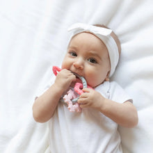 Load image into Gallery viewer, A baby in white outfit with a headband chews a Bumkins Silicone Teething Charms: Pink while laying on a white bedspread.
