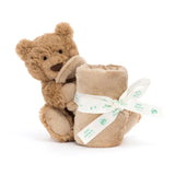 A soft brown Jellycat Soother Bartholomew Bear holding a rolled-up tan blanket tied with a printed ribbon.