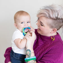 Load image into Gallery viewer, A smiling woman with short blonde hair wearing a purple sweater holds a baby chewing on Bumkins Silicone Teething Rings 4 Pack: Summer.
