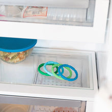 Load image into Gallery viewer, Bumkins Silicone Teething Rings 4 Pack: Summer sits on a glass refrigerator shelf, with a covered dish of food nearby.
