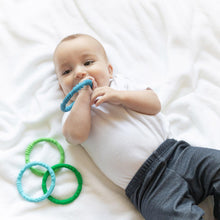 Load image into Gallery viewer, A baby on a white blanket, wearing a onesie and pants, chews a Bumkins Silicone Teething Rings 4 Pack: Summer while the others lay nearby.
