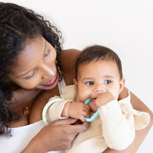 Load image into Gallery viewer, A smiling woman holds a baby, dressed in a beige shirt, chewing on a Bumkins Silicone Teething Rings 4 Pack: Spring.
