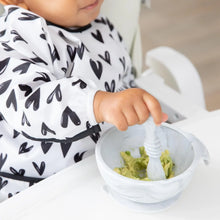 Load image into Gallery viewer, Close-up of a toddler in a Bumkins Sleeved Bib: Hearts eating mashed avocado with a spoon from a white bowl.
