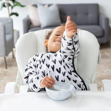 Load image into Gallery viewer, A toddler in a high chair wears a Bumkins Sleeved Bib: Hearts, giving a thumbs-up while holding a spoon by an empty bowl.
