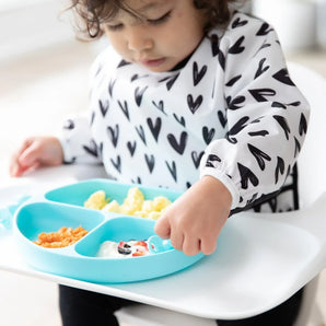 A toddler in a Bumkins Sleeved Bib: Hearts sits in a high chair, eating yogurt, fruit, & eggs from a blue divided plate.