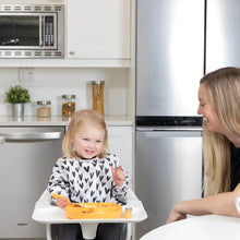 Load image into Gallery viewer, A toddler in a high chair wearing a Bumkins Sleeved Bib: Hearts holds a spoon over an orange plate, smiling at an adult in a modern kitchen.
