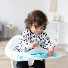 Load image into Gallery viewer, A toddler with curly hair wearing a Bumkins Sleeved Bib: Hearts sits in a high chair, removing a lid from a blue divided plate.
