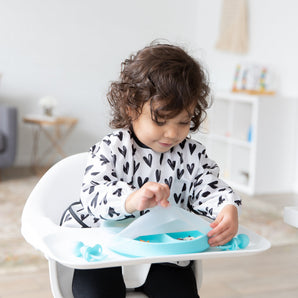 A girl wearing a black & white bib in a high chair lifts a Bumkins Silicone Stretch Lid for Grip Dish 3 Section, Grip Plate from a blue plate.
