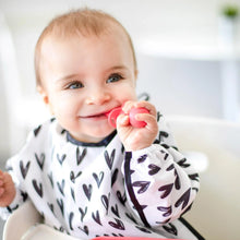 Load image into Gallery viewer, A happy baby in a Bumkins Sleeved Bib: Hearts chews a red toddler spoon in a bright room.

