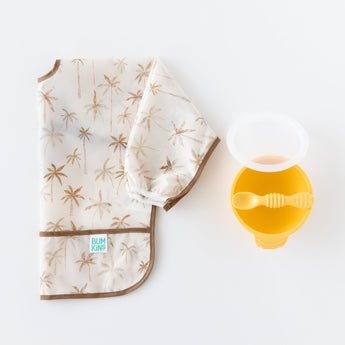 Bumkins Sleeved Bib: Palm Daze sits folded beside a yellow bowl and spoon on a white backdrop.