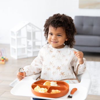 A toddler in a high chair wears a Bumkins Sleeved Bib: Palm Daze, holding a spoon and exploring baby-led weaning from a plate.
