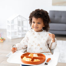 Load image into Gallery viewer, A toddler in a high chair wears a Bumkins Sleeved Bib: Palm Daze, holding a spoon and exploring baby-led weaning from a plate.
