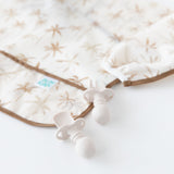 Close-up of Bumkins Sleeved Bib: Palm Daze next to two beige silicone utensils on a white background.