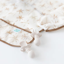 Load image into Gallery viewer, Close-up of Bumkins Sleeved Bib: Palm Daze next to two beige silicone utensils on a white background.
