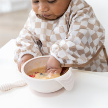 Load image into Gallery viewer, A baby wearing Bumkins Sleeved Bib: Sand Check eagerly explores porridge, embracing baby-led weaning at a white table.
