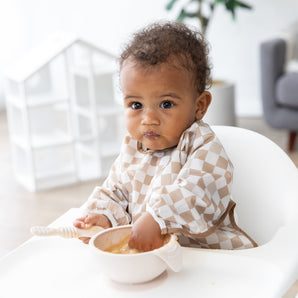 A baby in a high chair wears a Bumkins Sleeved Bib: Sand Check, trying baby-led weaning and wearing food cheerfully on their face.