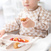 Load image into Gallery viewer, A toddler in a Bumkins Sleeved Bib: Sand Check enjoys baby-led weaning with fruit from a tan divided plate in a high chair.
