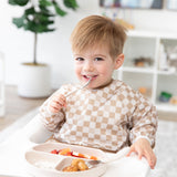 A happy toddler in a Bumkins Sleeved Bib: Sand Check sits in a high chair, enjoying baby-led weaning with a spoon from a beige plate.