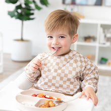 Load image into Gallery viewer, A happy toddler in a Bumkins Sleeved Bib: Sand Check sits in a high chair, enjoying baby-led weaning with a spoon from a beige plate.
