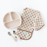 Bumkins Sleeved Bib: Sand Check is paired with a divided plate and matching spoon and fork on a white background.