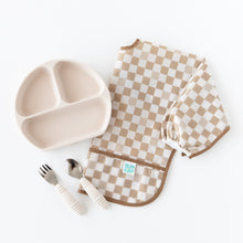 Load image into Gallery viewer, Bumkins Sleeved Bib: Sand Check is paired with a divided plate and matching spoon and fork on a white background.
