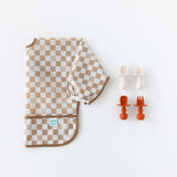 Bumkins Sleeved Bib: Sand Check lays folded next to two sets of silicone utensils on a white background.