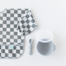 Load image into Gallery viewer, Bumkins Sleeved Bib: Charcoal Check lays next to a gray bowl and spoon on a white background.
