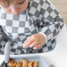 Load image into Gallery viewer, A child in a Bumkins Sleeved Bib: Charcoal Check reaches for pancakes, embracing baby-led weaning at a white table.
