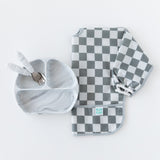 Bumkins Sleeved Bib: Charcoal Check is displayed with a gray divided plate and matching utensils on a white background.