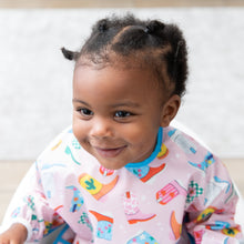 Load image into Gallery viewer, A girl with braided hair smiles, wearing a Bumkins Sleeved Bib: Boots with a blurred beige rug behind.
