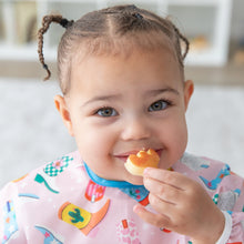Load image into Gallery viewer, Close-up of a smiling girl in pigtails wearing a Bumkins Sleeved Bib: Boots holding a snack to her mouth.
