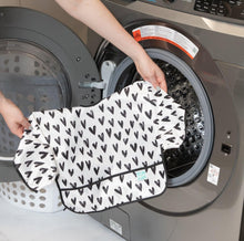 Load image into Gallery viewer, A person holds Bumkins Sleeved Bib: Hearts in front of an open front-loading washing machine.
